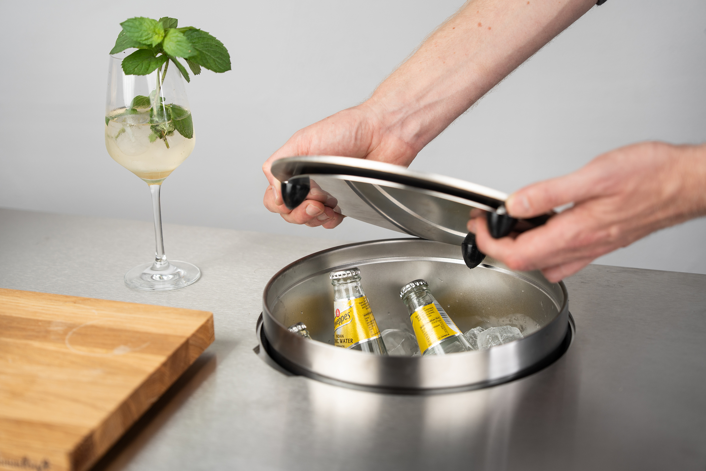 Einsetzbarer Edelstahldeckel wird über den integrierten Getränkekühler einer Flammkraft Outdoor-Küche gehoben, in dem Schweppes Tonic Water Flaschen auf Eis kühlen.