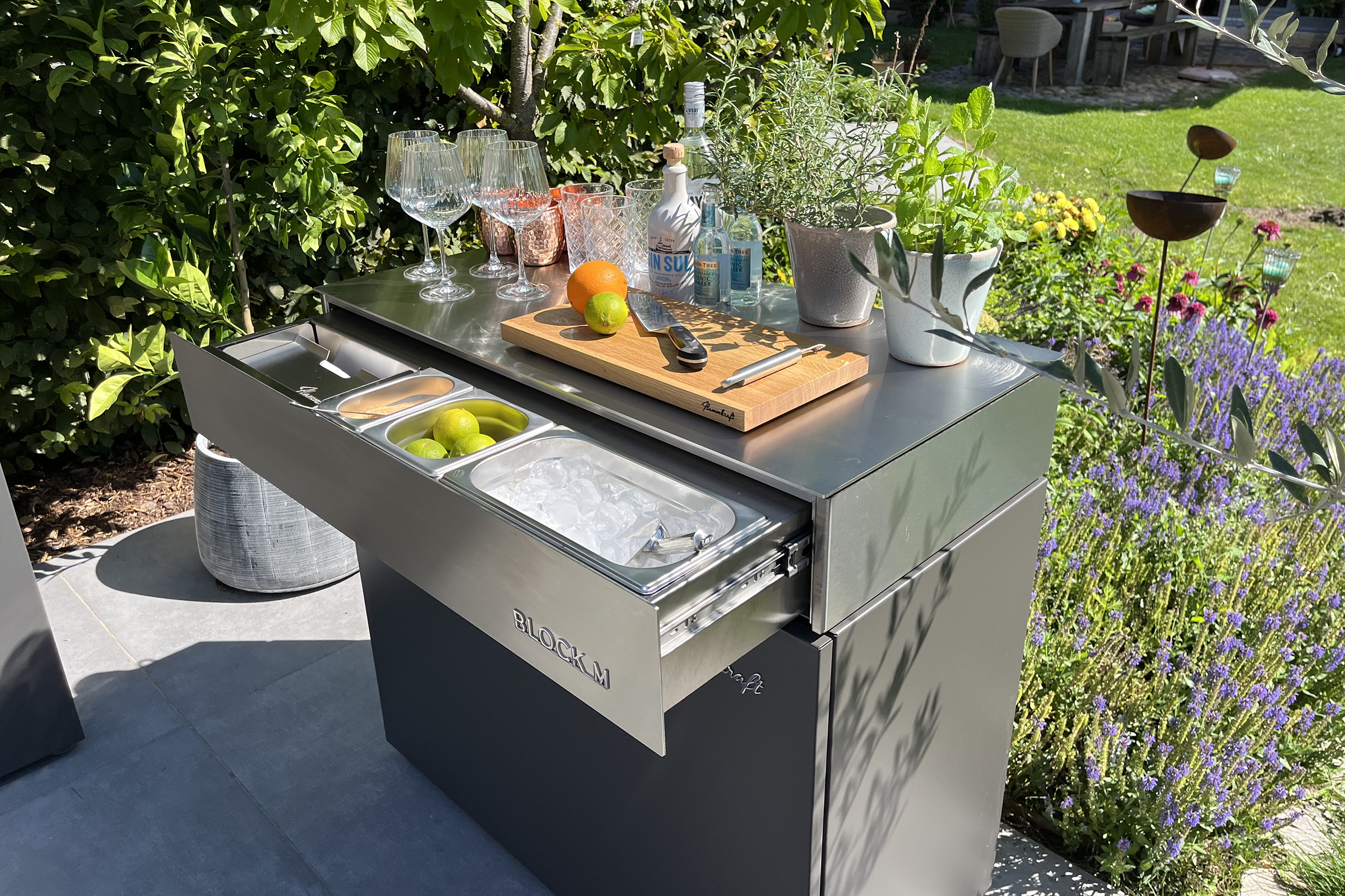 Flammkraft Block M Outdoor-Küchenmodul, im Garten als mobile Cocktailbar genutzt, mit ausgezogener Edelstahl-Schublade für Eiswürfel, Limetten, Gläser und Spirituosen auf der Arbeitsfläche.