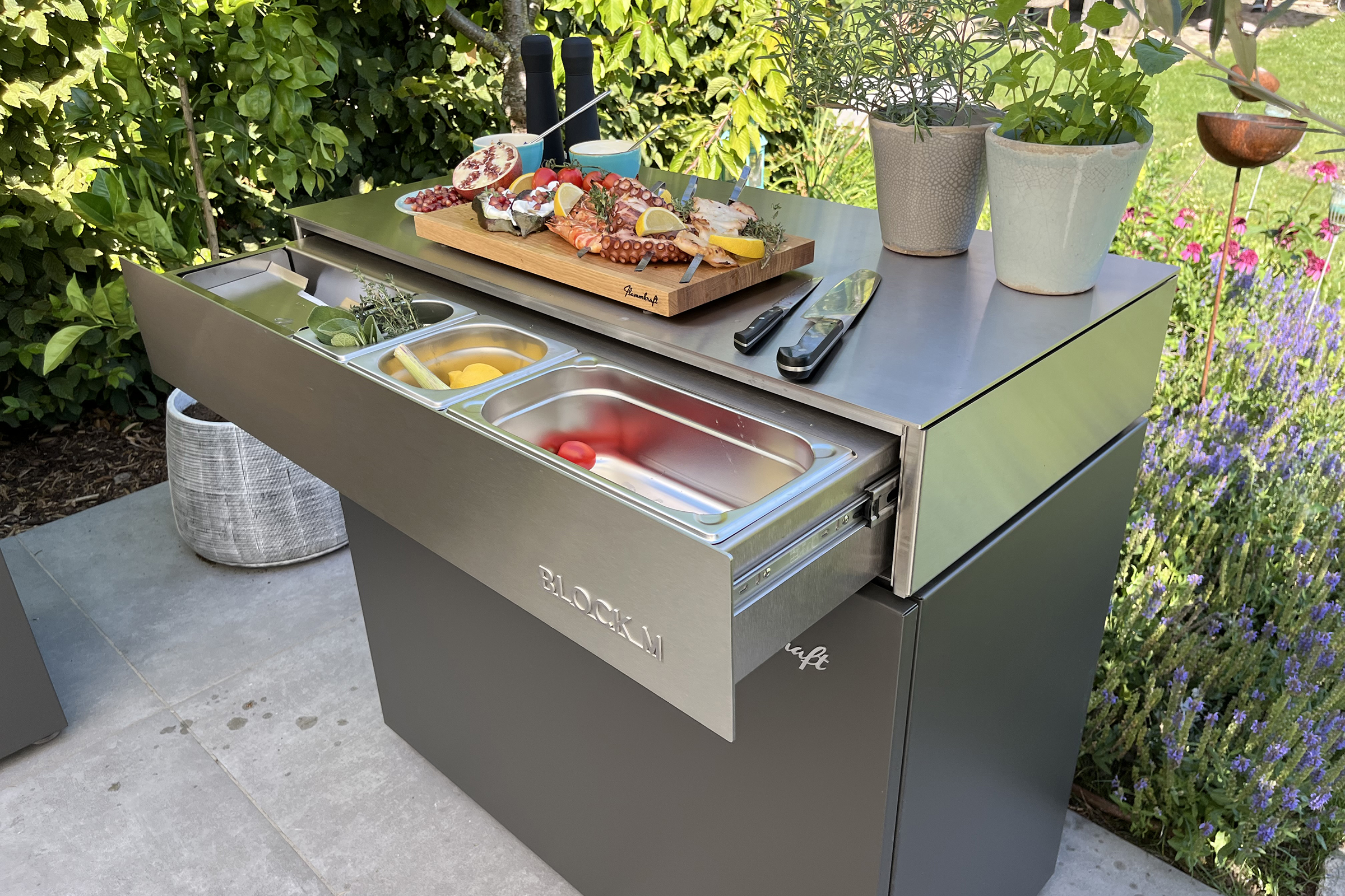 Flammkraft Block M Outdoor-Küchenmodul im Garten, vorbereitet für ein Meeresfrüchte-Grillen mit Oktopus, Garnelen und Kräutern auf einem Holzbrett sowie integrierter Gastronorm-Schublade.